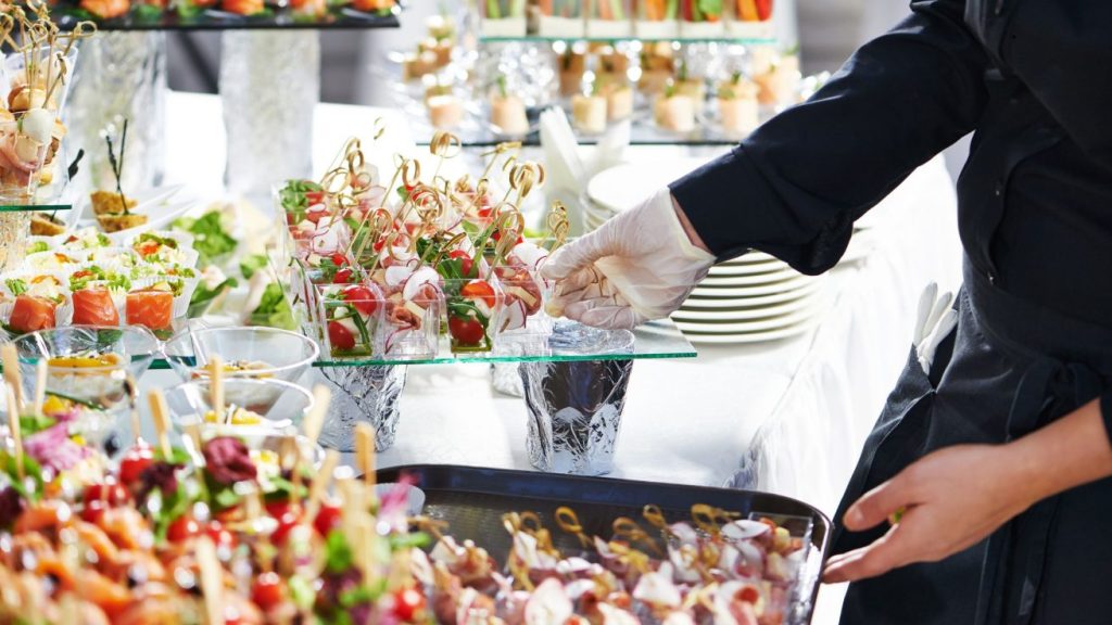 A-waiter-with-meat-dish-serving-catering-table-with-food-snacks-1310x737